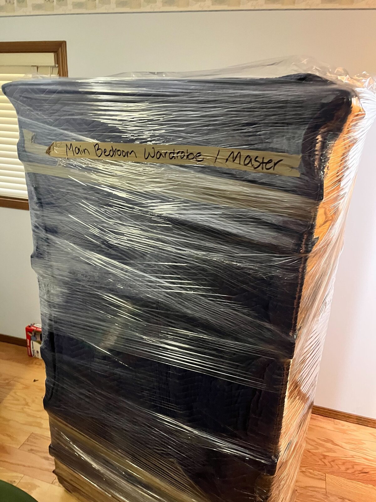 Tall wooden wardrobe protected with moving pads and stretch wrap and labeled for its destination room